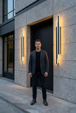 AI Studio Photo - Standing facing the camera, natural confident posture, arms relaxed at sides, front view, full body visible. CAMERA SHOT: A medium shot focusing on the wall light mounted on the exterior wall, showcasing its design and the warm illumination it casts. [Full body]